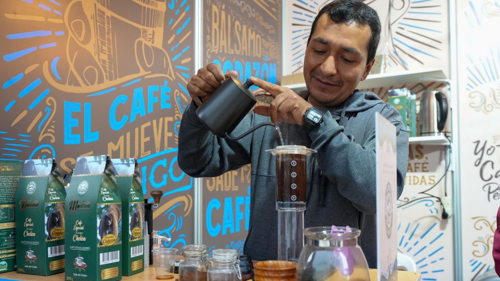 Un hombre prepara café vertiendo agua en un dispositivo de infusión, con empaques de café peruano variedad Cholón visibles en una exhibición. (DEVIDA)