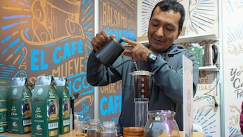 Hombre vierte agua caliente de una jarra negra sobre café molido en un infusor transparente. En el fondo, paquetes de café y un mural decorado con texto