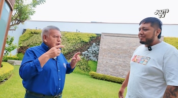 César Acuña, vestido con camisa azul, señala con la mano mientras habla con un joven con camiseta blanca en un jardín con paredes de follaje verde