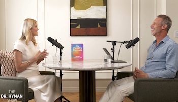 Mujer y hombre sentados a una mesa redonda con dos micrófonos, dos vasos de agua, una tableta y el libro 'The Fertility Formula'. Logo 'The Dr. Hyman Show'