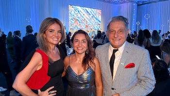 Tres personas sonríen y posan en un evento formal con fondo azul. Dos mujeres vestidas elegantemente y un hombre con saco a cuadros y pañuelo rojo