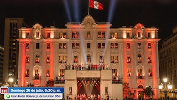 El Gran Hotel Bolívar de Lima iluminado de rojo y blanco con una bandera peruana en la parte superior, personas en ventanas, un escenario con músicos y público