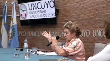 Una mujer mayor con cabello rubio peinado y blusa rosa floral habla en un micrófono. Se ven banderas, un logo de Uncuyo y una pared de ladrillo al fondo