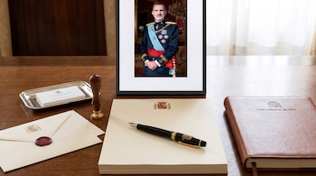 Una mesa de madera con papelería oficial de la Casa Real española (sobres, papel, pluma, cuaderno) y un retrato enmarcado del Rey Felipe VI.