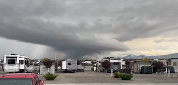 Grandes nubes oscuras de tormenta cubren el cielo detrás de un estacionamiento con casas rodantes, vehículos y montañas al fondo