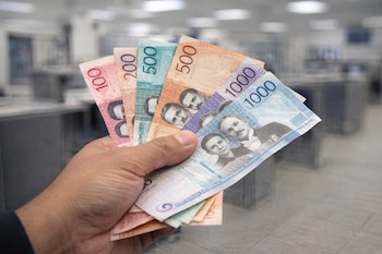 Primer plano de una mano sosteniendo billetes de pesos dominicanos de 100, 200, 500 y 1000, con un fondo difuminado de un espacio interior tipo banco.