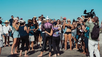 Una persona de espaldas con traje de neopreno y gorro de natación sobre la arena, frente a varias personas con cámaras de foto y video