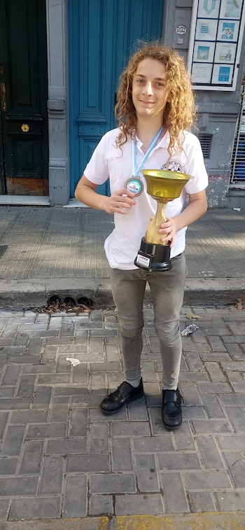Un joven de cabello rizado con una camiseta blanca sostiene un trofeo dorado y una medalla en una acera de adoquines frente a puertas