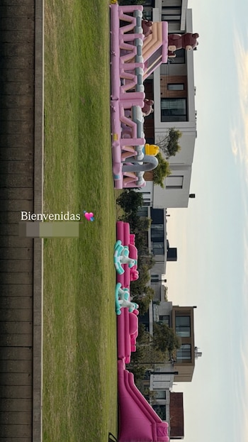 Césped verde, varios juegos inflables rosas y celestes con toboganes y figuras de osos. Casas modernas en el fondo, cielo azul claro. Texto: Bienvenidas