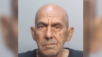 Fotografía policial de un hombre calvo de edad avanzada con barba corta y bigote, mirando directamente a la cámara sobre un fondo liso