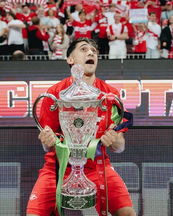 Un hombre joven con camiseta roja sostiene un trofeo de cristal roto con la tapa ladeada, con expresión asombrada y mirando hacia arriba