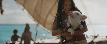 Moana sonriente abraza a un cerdito blanco con el hocico hacia arriba, con el mar turquesa y un velero borroso de fondo