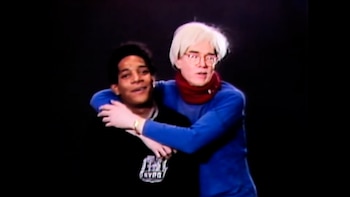 Andy Warhol, de cabello rubio platino y gafas, abraza por la espalda a Jean-Michel Basquiat, de cabello oscuro, sobre un fondo negro