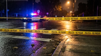 Cinta amarilla con la leyenda Policía no pasar en primer plano sobre asfalto mojado y un patrullero con luces encendidas al fondo.