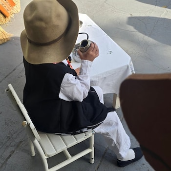 Vista trasera de una persona con sombrero, camisa blanca y chaleco negro. Está sentada en una silla blanca, sosteniendo un mate y un termo sobre una mesa blanca