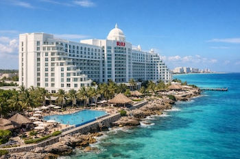 Vista aérea lateral del Hotel Riu Palace de Cancún, un gran edificio blanco con cúpula, rodeado de piscinas, palmeras y la costa rocosa junto al mar turquesa.