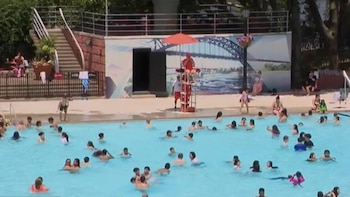 Piscina exterior azul llena de personas nadando. Un socorrista en uniforme rojo está en su torre con sombrilla. Un mural de un puente decora el fondo