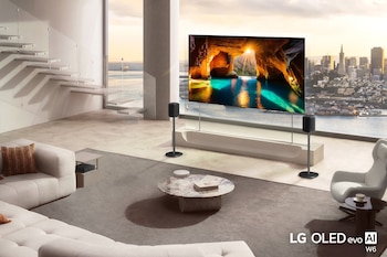 Pantalla delgada de televisor entre dos altavoces en una sala moderna con sofá, mesa de mármol y ventanal hacia un paisaje urbano