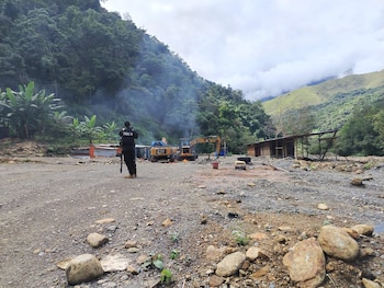 Oficial de policía observa dos excavadoras amarillas y campamento rústico en un área montañosa boscosa con humo y tierra