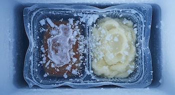 Bandeja plástica con pastel de carne y puré de papas congelados, cubiertos con cristales de hielo, dentro de un congelador.