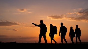 Siluetas de cinco personas caminando sobre una loma al atardecer; el cielo muestra tonos naranjas, amarillos y púrpuras con nubes; uno de ellos señala hacia adelante.