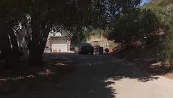 Casa con garaje blanco y detalles de piedra, rodeada de árboles frondosos y follaje, con un coche oscuro en el camino de entrada bajo la luz del sol