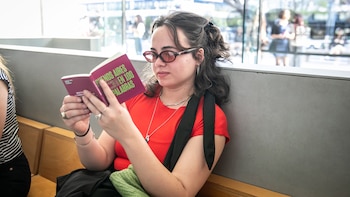 Una mujer joven con cabello oscuro y lentes marrón-rojizos lee un libro pequeño con la cubierta fucsia que dice 'BUENOS AIRES EN 100 PALABRAS'