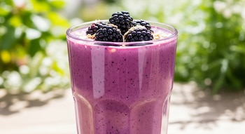 Licuado cremoso de color violeta intenso en un vaso alto, adornado con moras frescas y avena espolvoreada, sobre una superficie clara con fondo verde difuminado.