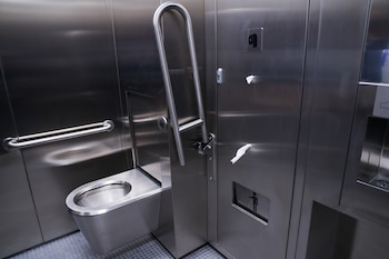 Un baño público inteligente de acero inoxidable con inodoro, barras de apoyo, y dispensadores de papel en un espacio moderno y metálico