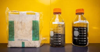 Un paquete de baterías LFP, un frasco con polvo de LFP agotado y un frasco con polvo de LMFP reciclado sobre una superficie oscura