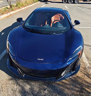 Vista frontal de un coche deportivo McLaren de color azul oscuro, estacionado en un área pavimentada con reflejos de sol