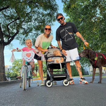 Muni Seligmann sonríe junto a su esposo, hija en bicicleta, bebé en cochecito y perro en un sendero arbolado, bajo la luz del sol