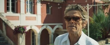 Hombre con gafas de sol y camisa blanca frente a una casa de dos pisos de color terracota con palmeras y arbustos verdes al costado