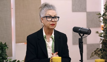 Mujer con cabello gris, gafas y labial rojo, vestida con blazer negro y camisa clara, sentada frente a un micrófono de estudio y una taza amarilla