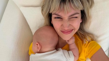 Úrsula Corberó, con cabello rubio y flequillo, guiña un ojo mientras sostiene a su bebé Vito Darín, cuya nuca y oreja son visibles