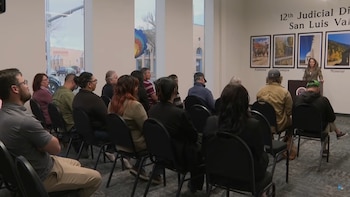 Una mujer rubia habla desde un podio marrón en una sala con una audiencia sentada; en la pared posterior se lee "12th Judicial District San Luis Valley"