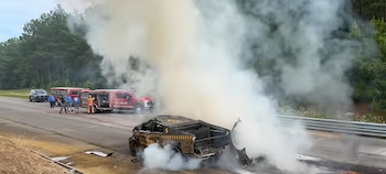 Coche en llamas en la carretera con humo denso. Dos camiones de bomberos, una camioneta policial y personal de emergencia visibles cerca. Árboles al fondo