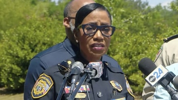 Cerelyn J. Davis, jefa de policía, habla en un podio con micrófonos. Viste un uniforme azul con insignias y gafas. Fondo verde borroso
