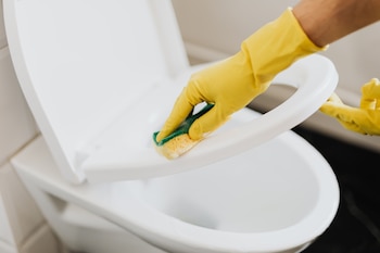 Mano con guante amarillo limpiando la tapa de un inodoro blanco con una esponja de dos colores