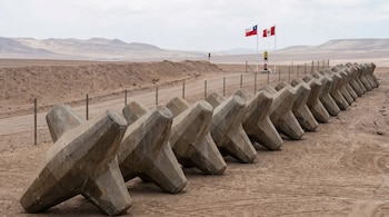 Larga fila de bloques de concreto en forma de cruz, o tetrápodos, sobre terreno arenoso de desierto. Al fondo, un hito fronterizo con banderas de Chile y Perú.