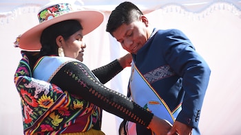 Una mujer con sombrero y vestimenta tradicional de colores vivos ajusta la banda azul y tricolor a Leonardo Loza, quien viste un traje oscuro