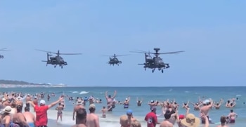 Cuatro helicópteros militares vuelan bajo sobre una playa con numerosas personas en el agua y la arena bajo un cielo azul despejado