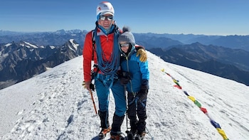 Un adulto y un niño con ropa de montaña, cascos y gafas de sol posan en una cima nevada junto a una tira de banderas de colores