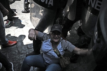 Una mujer con gorra, gafas, camiseta clara y jeans está sentada en el suelo, rodeada por piernas y escudos transparentes que apenas revelan siluetas detrás