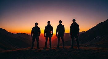 Cuatro siluetas de hombres de pie en fila, con gorros y chaquetas, sobre una colina. El sol poniente detrás de ellos ilumina un paisaje de montañas.