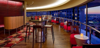 Lounge de lujo con grandes ventanales curvos, vistas urbanas nocturnas. Muebles rojos, mesas con velas, taburetes de madera, mostrador y persiana