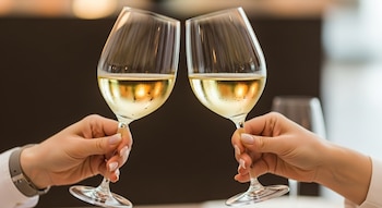 Dos manos brindan con copas de vino blanco llenas, con condensación en el cristal, sobre un fondo borroso.