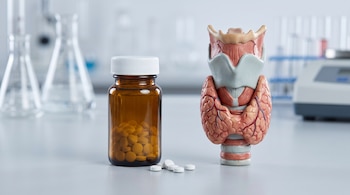 Un frasco de vidrio oscuro con pastillas, varios comprimidos sueltos y una maqueta anatómica de la tiroides sobre una mesa de laboratorio.