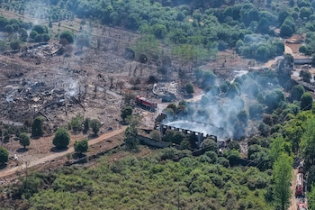Vista aérea de un área devastada por una explosión, mostrando edificios destruidos, escombros, humo blanco y camiones de bomberos en una zona arbolada