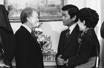 Fotografía en blanco y negro de tres personas. Dos hombres conversan de perfil y una mujer los observa. Al fondo, un cuadro con barcos y arreglo floral